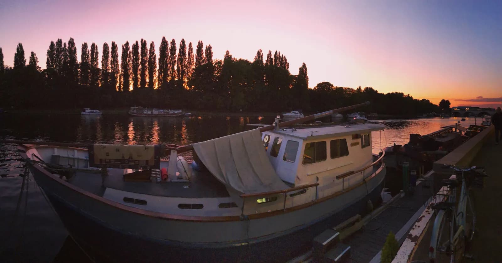 A small boat docked on a river at sunset, with a line of trees silhouetted against the pink and orange sky.
